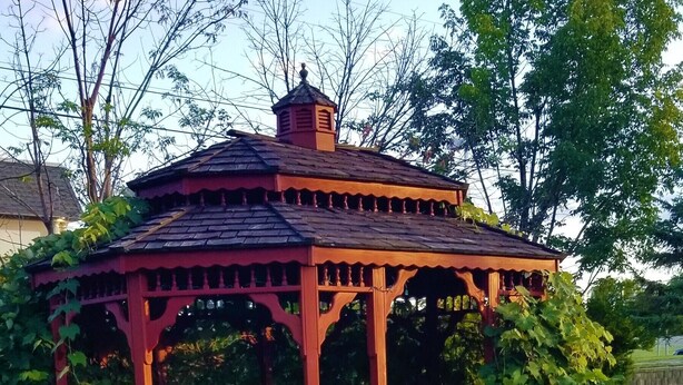 A wooden gazebo with a tiled roof, surrounded by greenery and a picnic table in a garden setting. - Lake Erie Lodge (Erie)