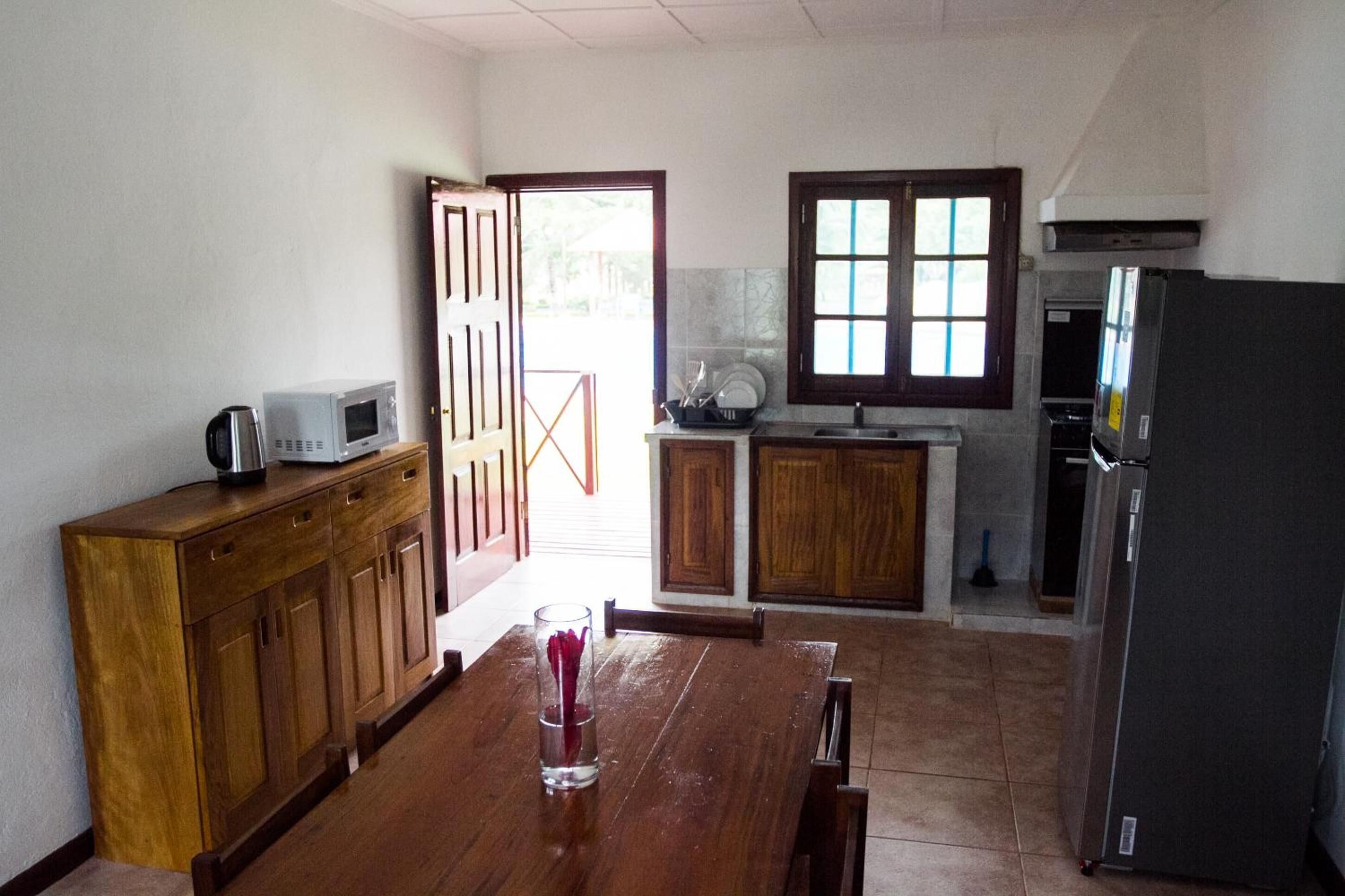Una cocina con armarios de madera, microondas, refrigerador y ventana con persianas.