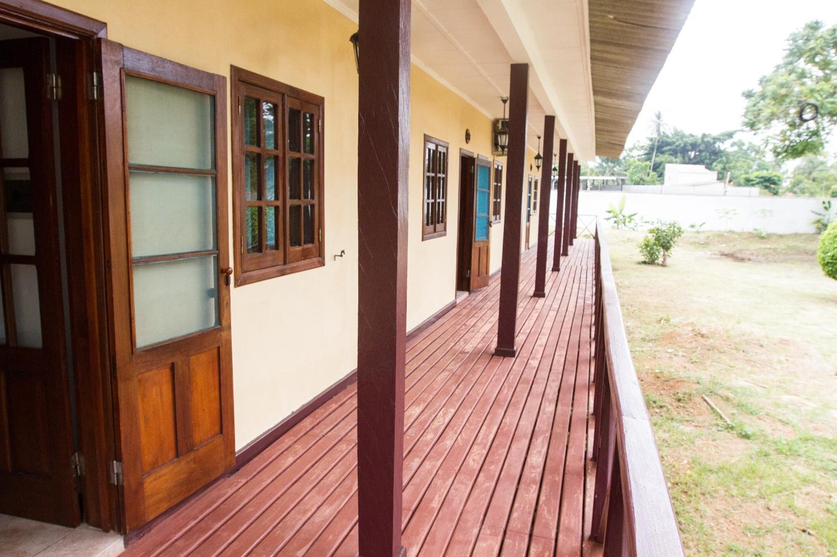 A covered wooden deck with a railing, a yellow wall with multiple windows, and a grassy area visible in the background.