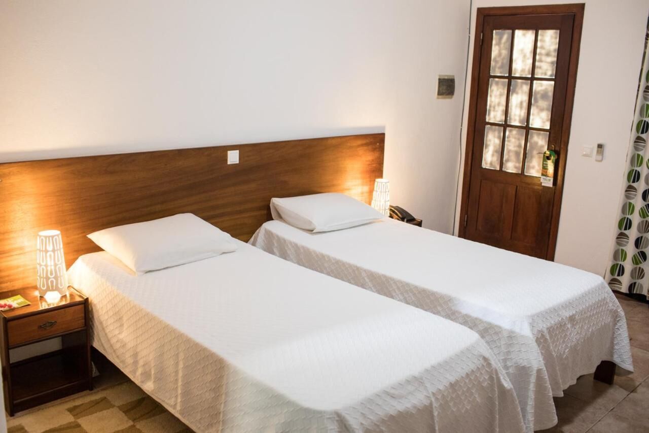 Una habitación con dos camas individuales, cabeceros de madera, ropa de cama blanca, una mesita de noche con lámpara y una puerta con ventana.