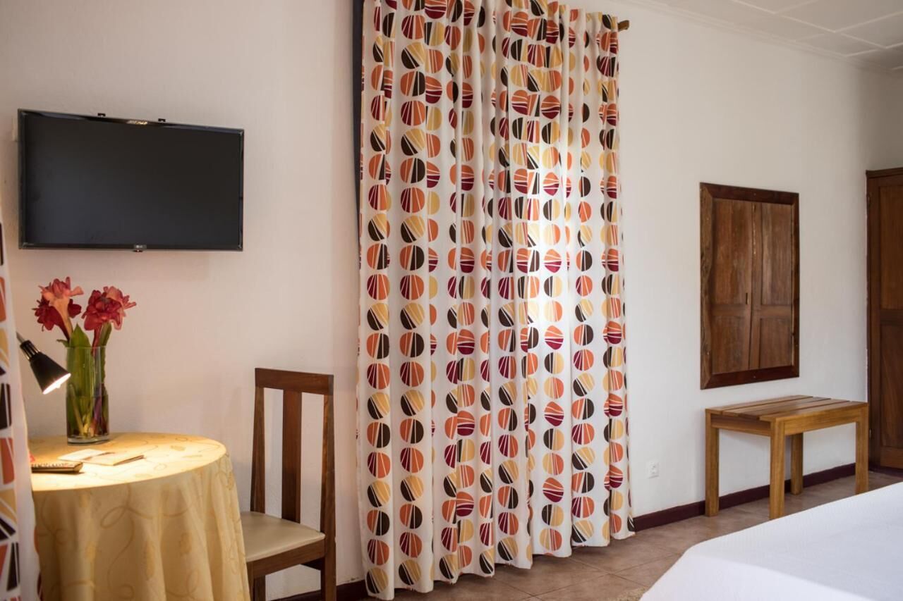 A room with a bed, a wooden table with a lamp, a chair, a TV mounted on the wall, and a curtain with a circular pattern.