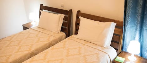 Dos camas individuales con cabeceros de madera, una mesita de noche con lámpara y una cortina azul estampada.