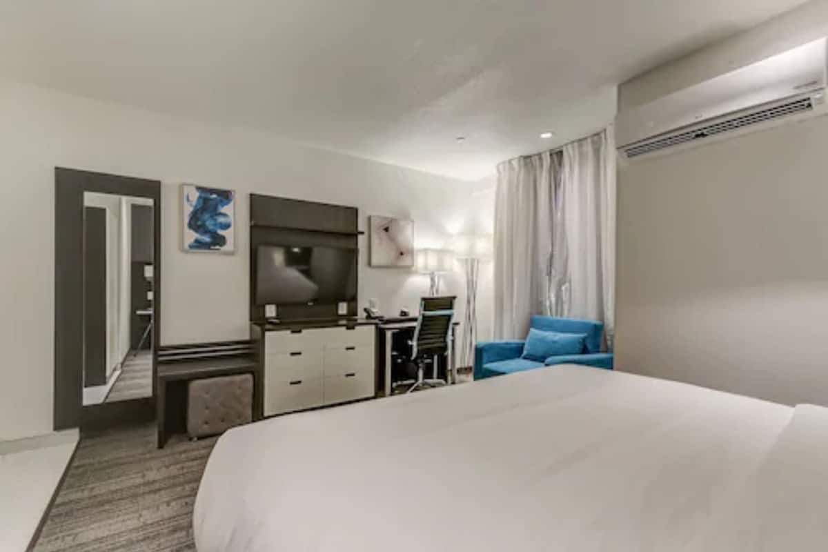 A hotel room with a large bed, a desk with a chair, a mirror, a lamp, and a blue armchair.