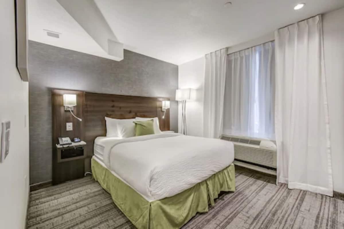 A hotel room with a large bed, a nightstand, a wall-mounted phone, and a window with curtains.