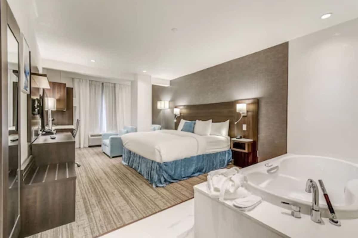 A modern hotel room with a large bed, a separate sitting area, a bathroom with a bathtub, and a desk with a chair.