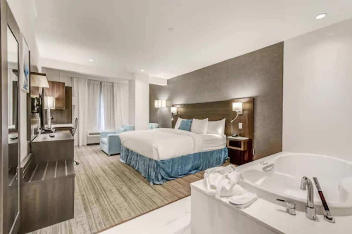 A modern hotel room with a large bed, a separate sitting area, a bathroom with a bathtub, and a desk with a chair.