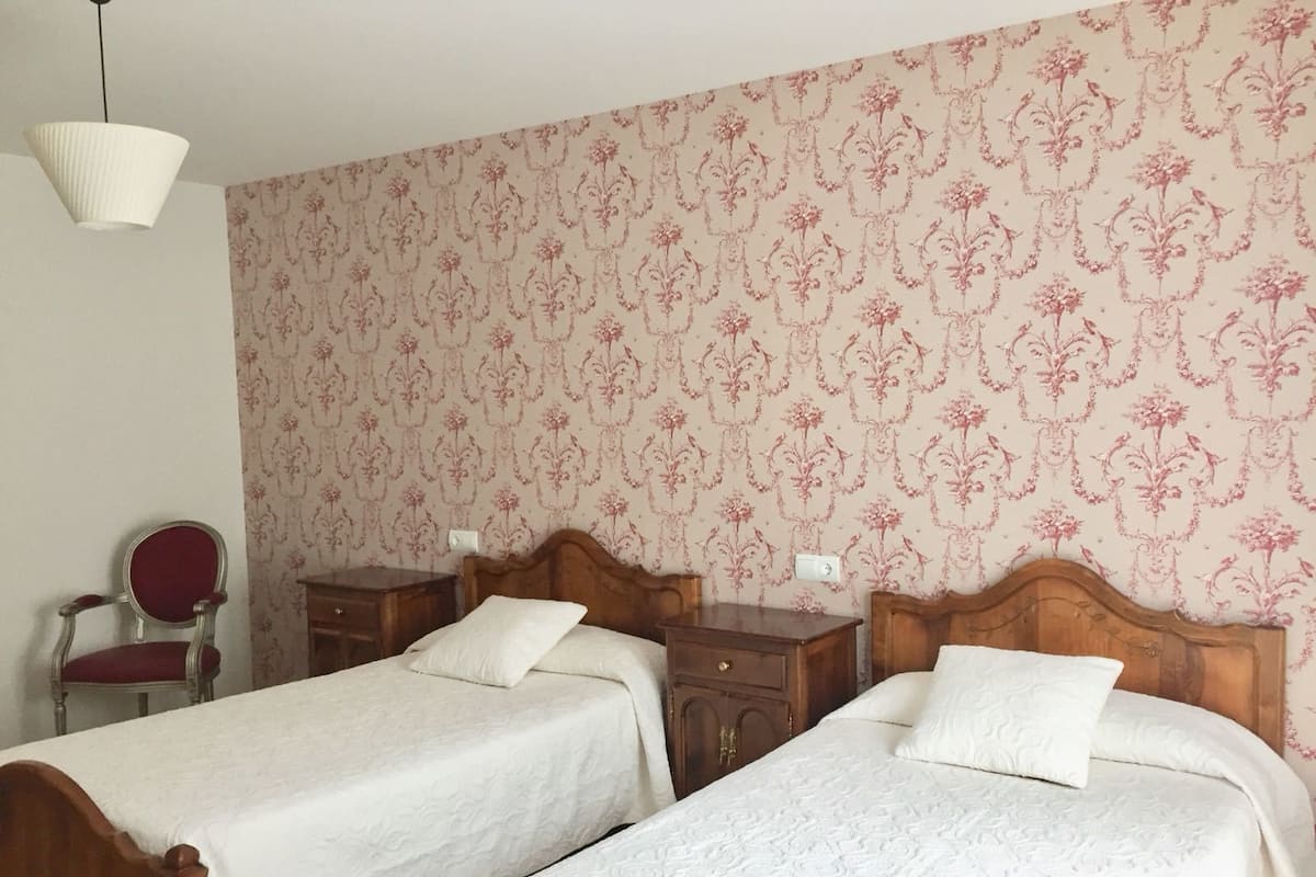 A bedroom with two single beds, a wooden nightstand, a chair, and a patterned wallpaper.
