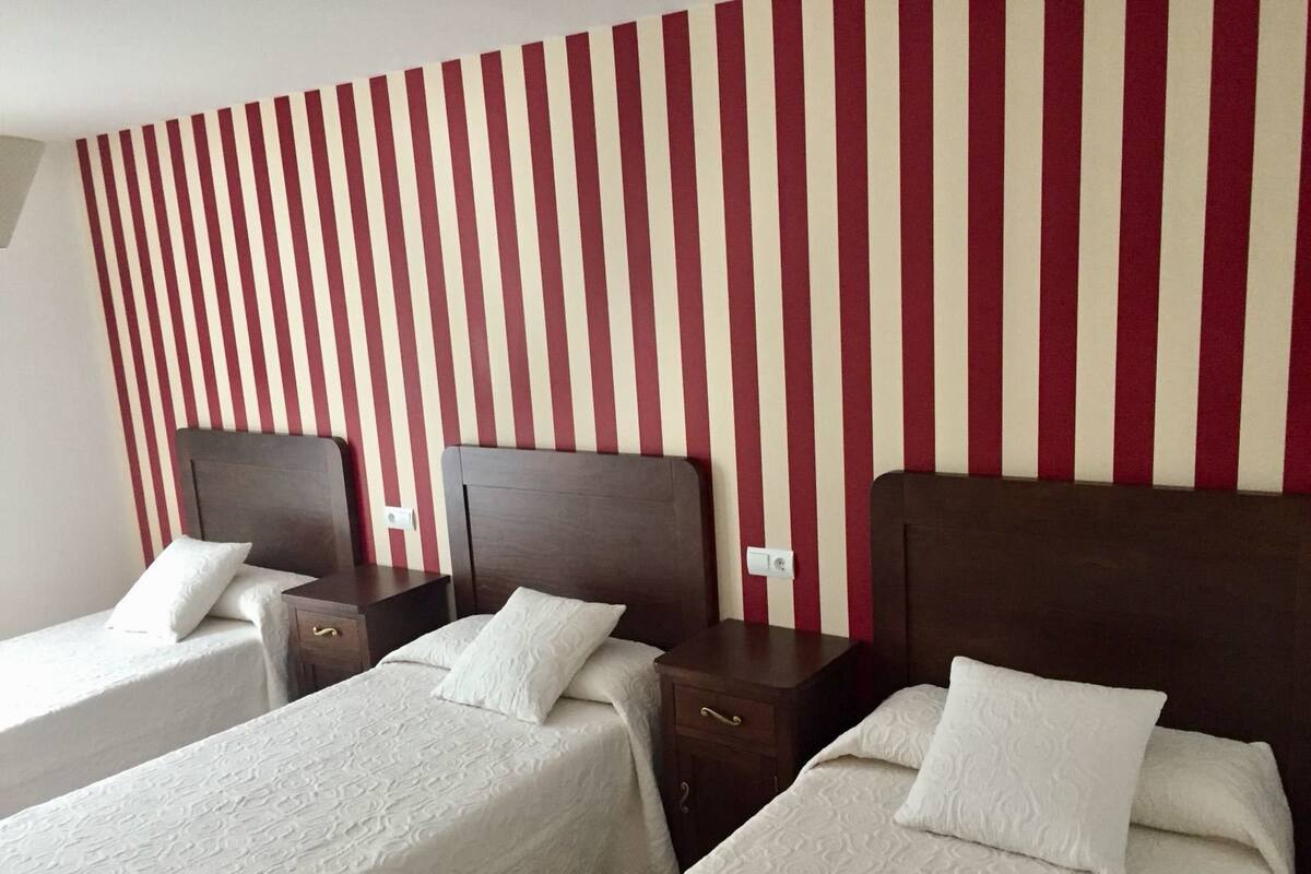 A hotel room with three single beds, each with a wooden headboard and white bedding. The wall features vertical red and white stripes.