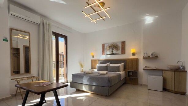 A modern hotel room with a bed, bedside table, and a small kitchenette. - Deluxe Suites (Chania)