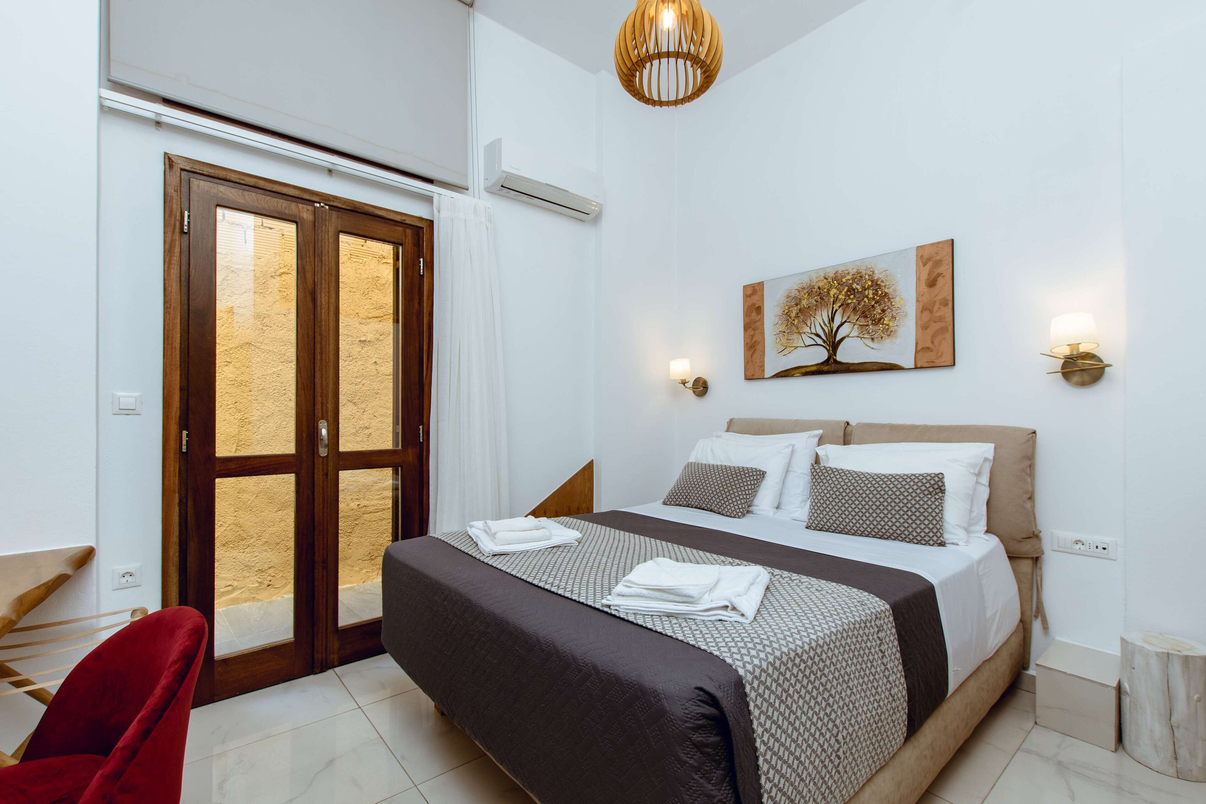 A bedroom with a bed, a red chair, a wooden door, and a wall-mounted painting.