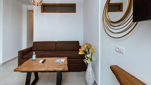 A modern living room with a brown sofa, a wooden coffee table, a vase with flowers, a wall-mounted mirror, and a ceiling light fixture. - Deluxe Suites (Chania)