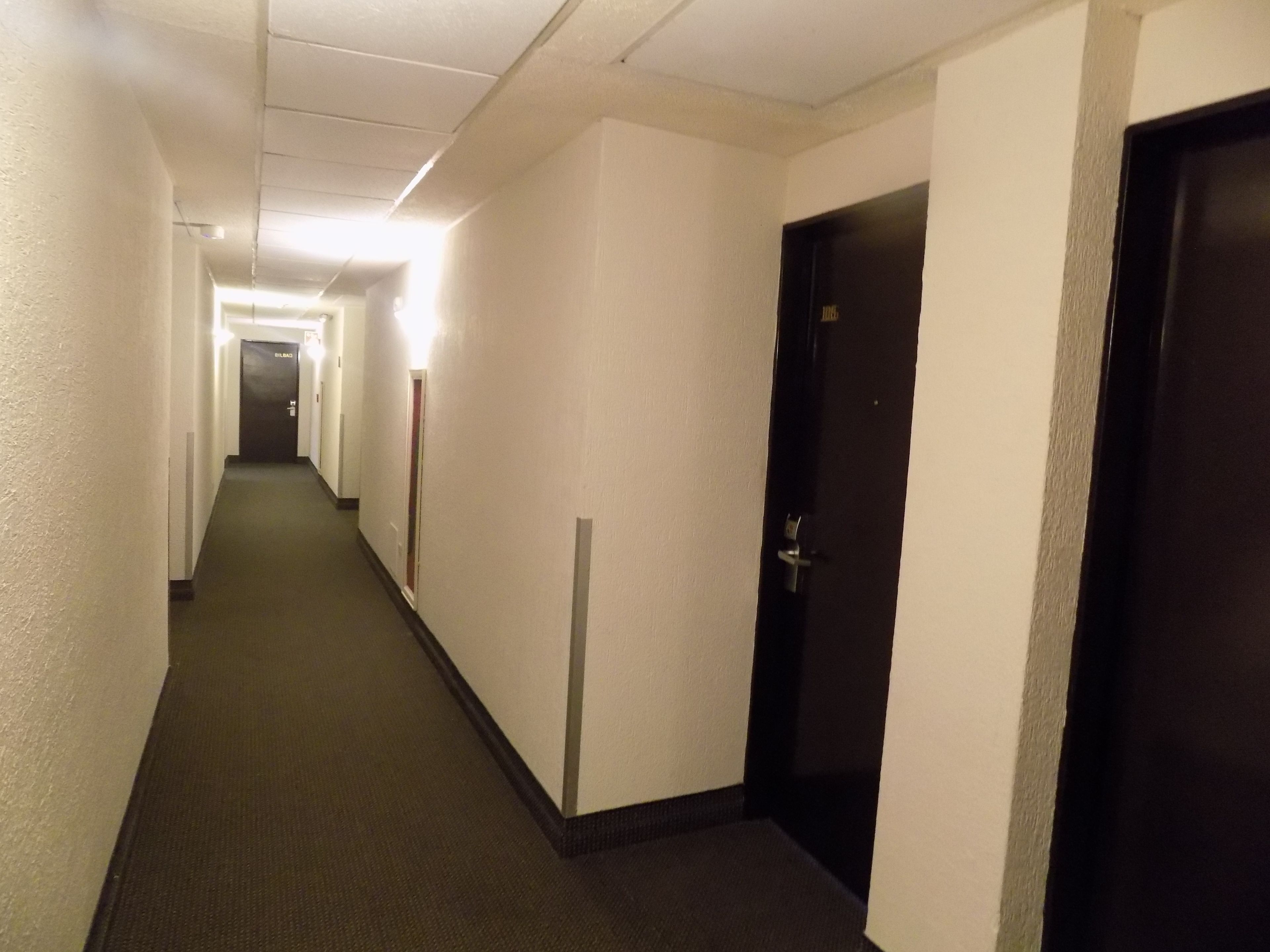 Corredor de hotel comprido e estreito, com paredes brancas e piso coberto de carpete, que leva a várias portas.