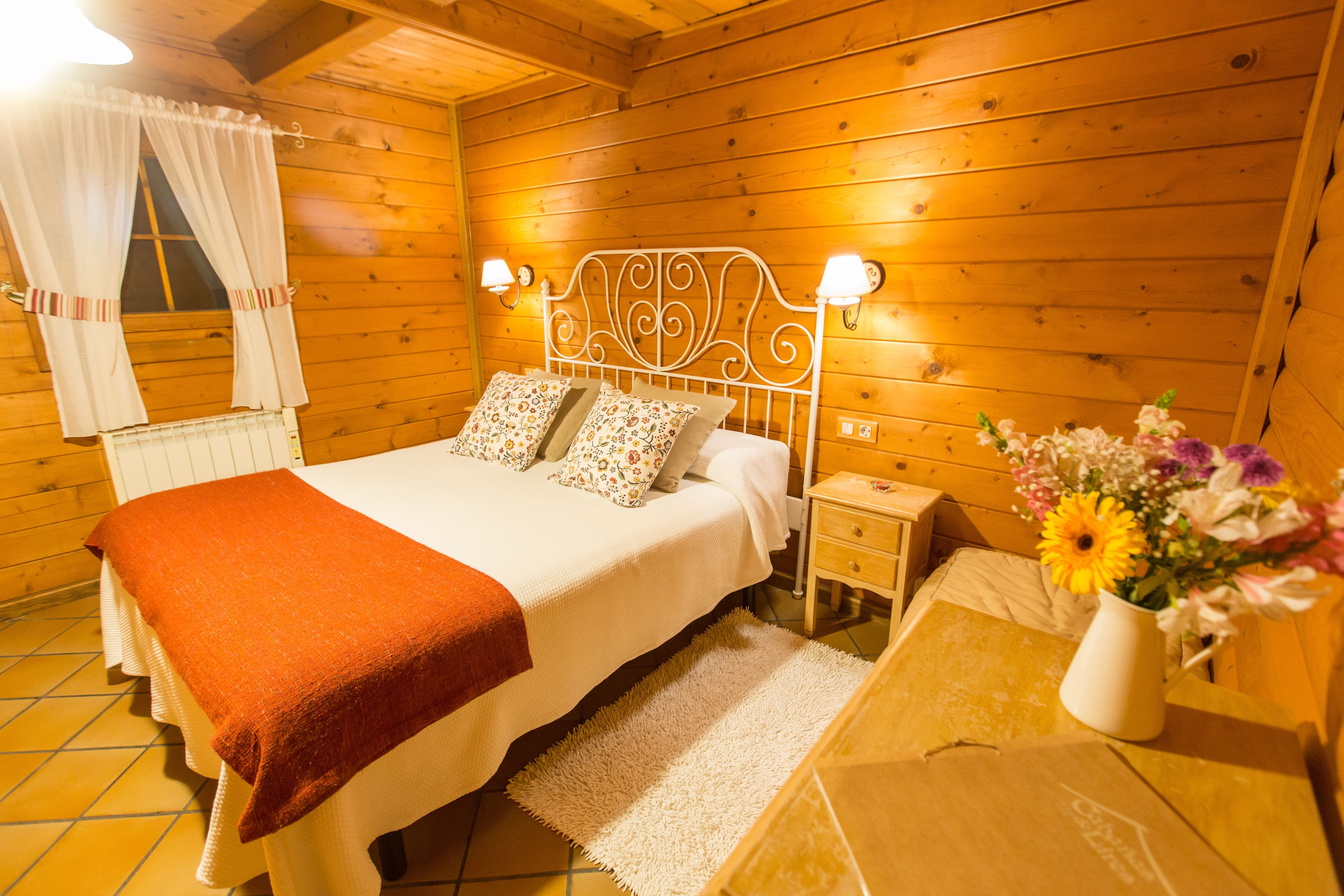 Un dormitorio de cabaña de madera con una cama, mesitas de noche, un florero con flores y una ventana con cortinas.
