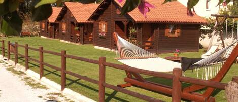 A wooden cabin-style building with a red-tiled roof, surrounded by a wooden fence, and featuring a hammock and a table with flowers.