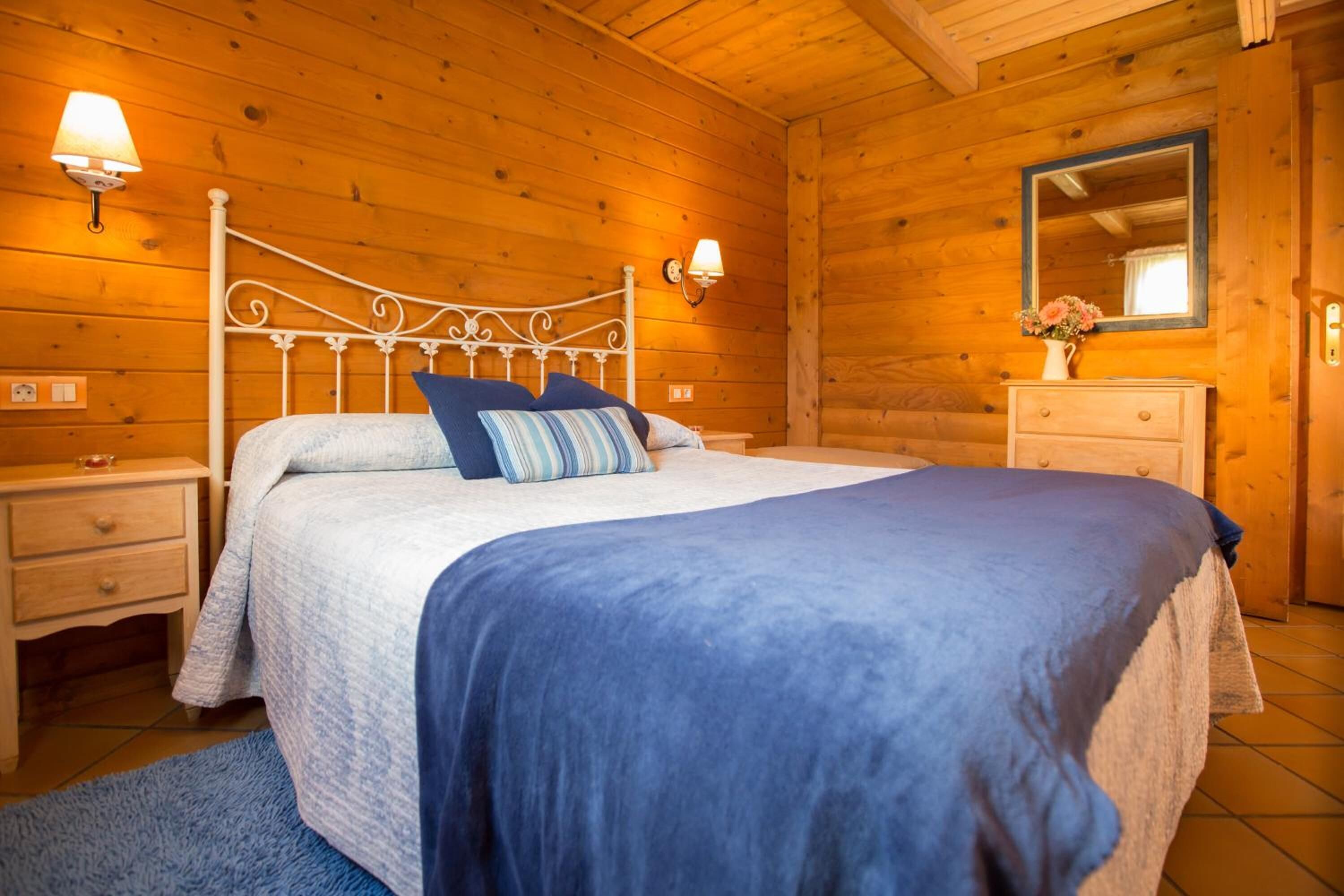 Un dormitorio de cabaña de madera con una cama, mesitas de noche, un aparador, un espejo y apliques de pared.