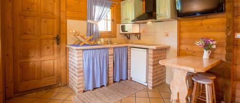 Interior de uma cabana de madeira com cozinha, incluindo micro-ondas, pia e uma pequena geladeira. Há um balcão de madeira com banquetas e uma televisão fixada na parede.