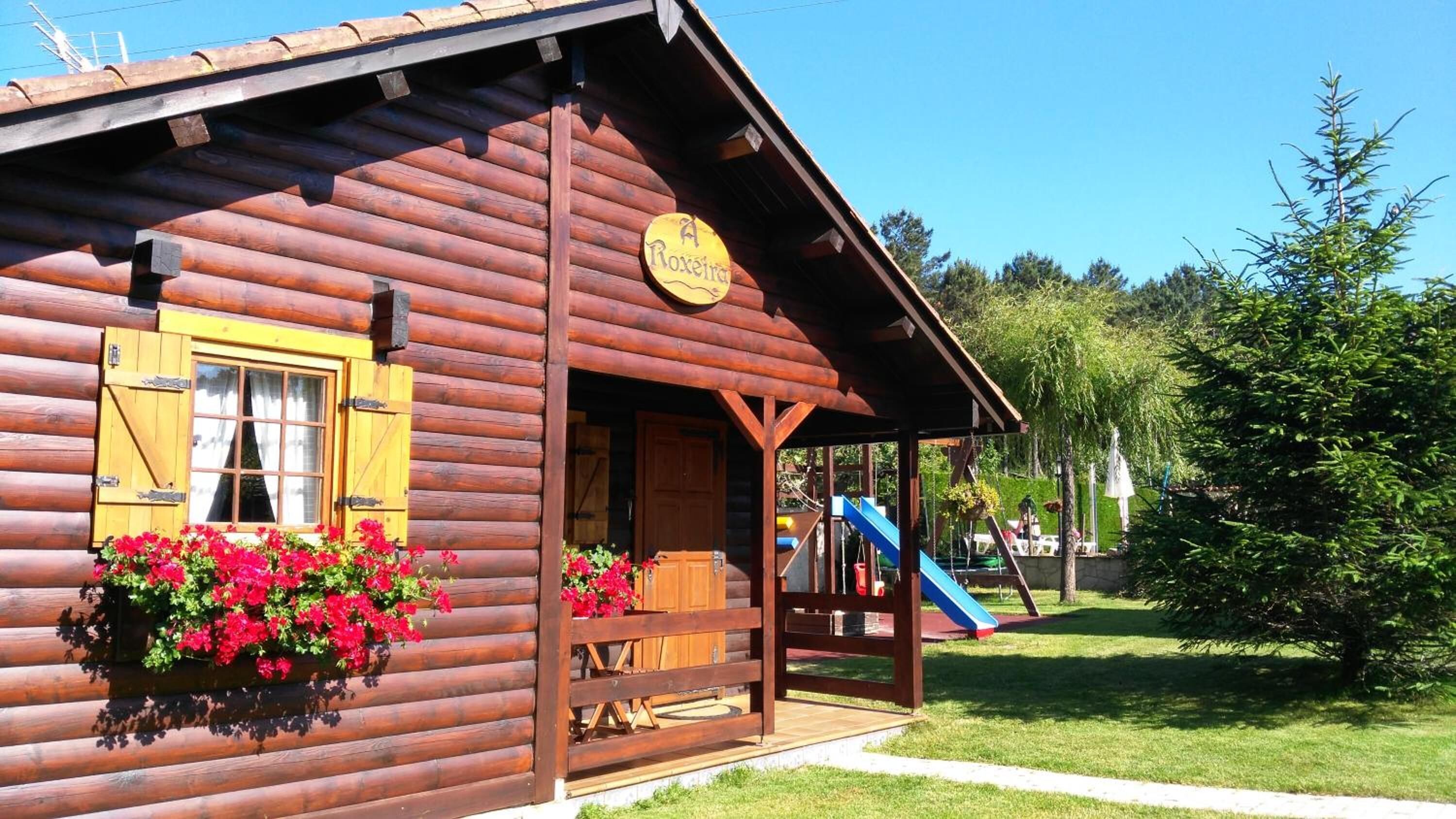 Une cabane en bois avec un porche, une enseigne indiquant «A Roxerra», et un toboggan bleu en arrière-plan.