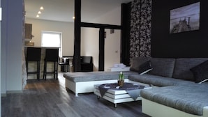 A modern living room with a grey sofa, a white coffee table, and a small side table. There is a black chair and a desk in the background. - Hotel Villa Alice (Thale)