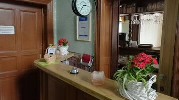 Un área de recepción con mostrador de madera, un reloj, un cartel con el horario del restaurante y una estantería con cristalería.