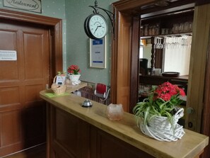A reception area with a wooden counter, a clock, a sign indicating restaurant hours, and a shelf with glassware. - Hotel Villa Alice (Thale)