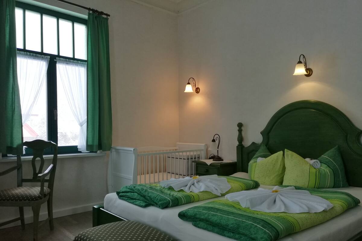 A bedroom with a green headboard, a white mattress, green and white bedding, a wooden chair, a nightstand with a lamp, and a window with green curtains.