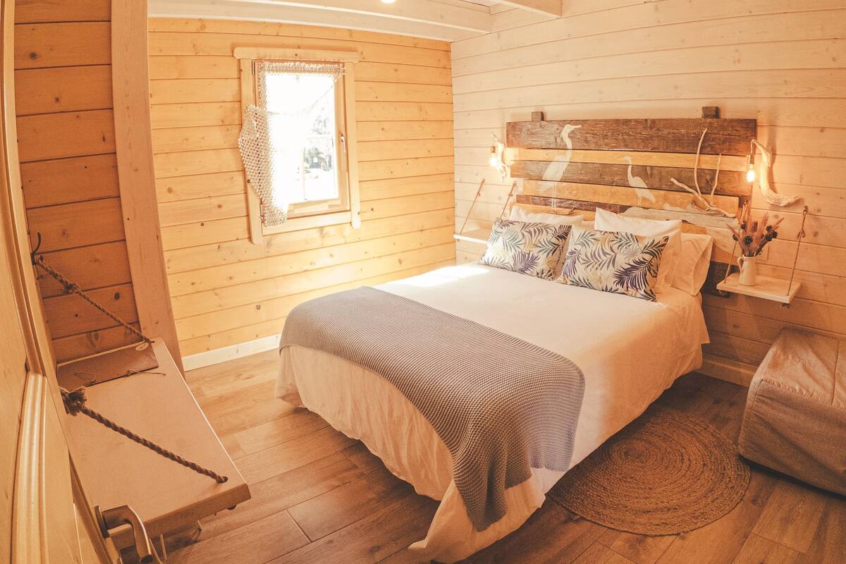 A cozy bedroom with a wooden ceiling, a bed with a headboard, a window, and a bedside table.