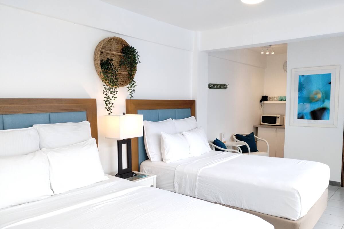 Een hotelkamer met twee bedden, een nachtkastje met een lamp, een zithoekje met een stoel, en een magnetron op een aanrecht.