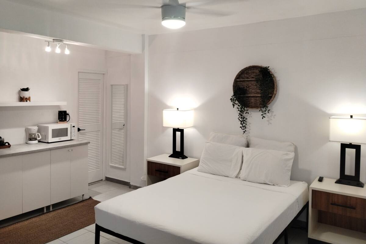 Een slaapkamer met een bed, nachtkastjes, een lamp, een plafondventilator, een magnetron en een koffiezetapparaat op een aanrecht.