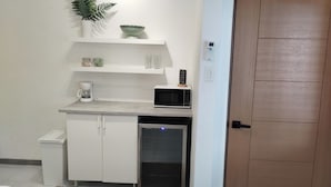 Eine kompakte Küchenzeile mit Mikrowelle, Kaffeemaschine und kleinem Kühlschrank. Weiße Schränke und eine Arbeitsplatte sind vorhanden.