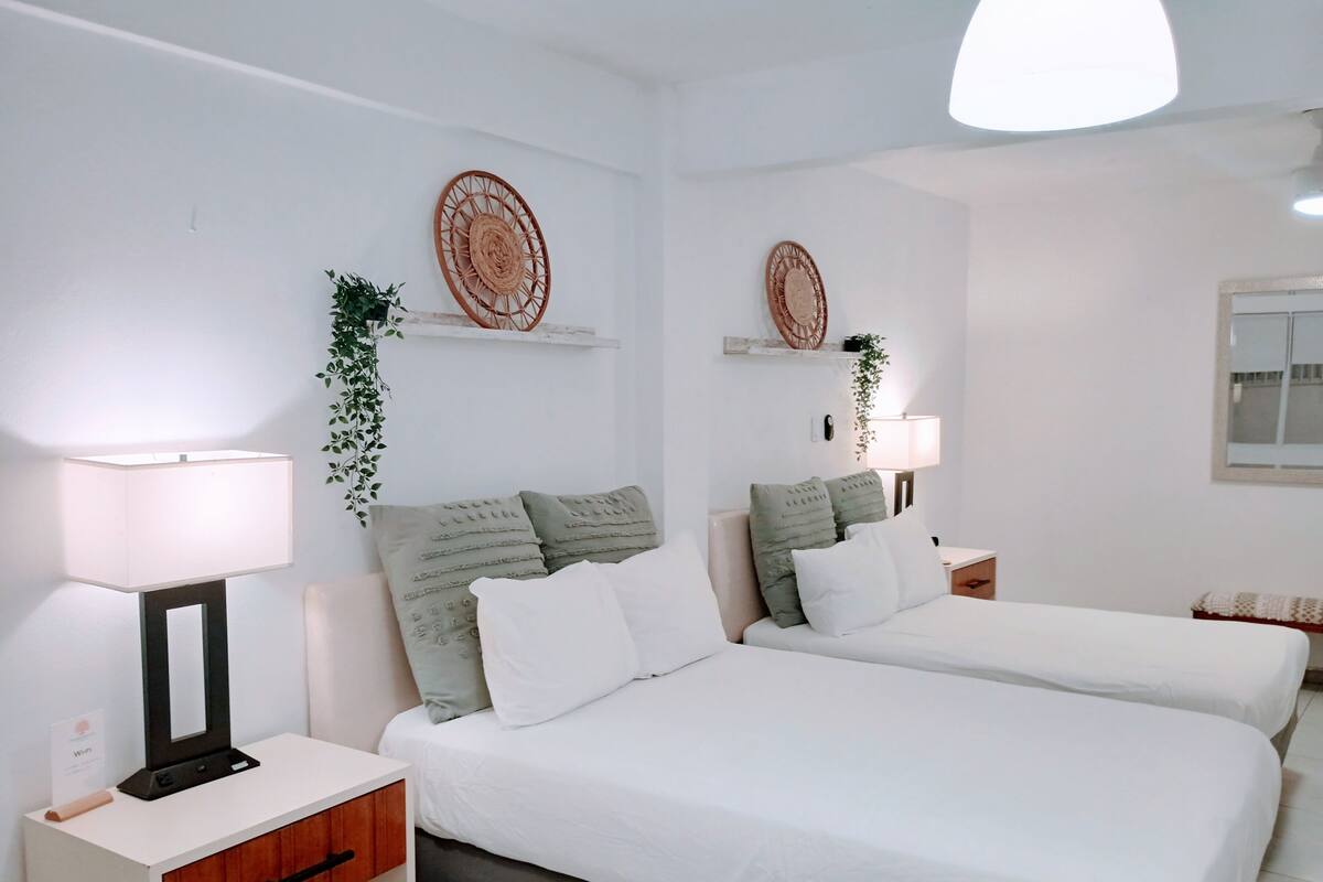 Een hotelkamer met twee bedden, wit bedlinnen, een nachtkastje met een lamp, een klein houten kastje en decoratieve wandkunst.