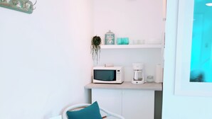 Eine kompakte Küchenzeile mit Mikrowelle, Kaffeemaschine und einer kleinen Ablage. Ein weißer Stuhl mit türkisnem Sitzkissen und ein kleiner Tisch mit einer Karte darauf.