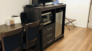 Un espace de vie compact comprenant un bureau en bois foncé, un téléviseur à écran plat, un four à micro-ondes et un petit réfrigérateur. Il y a deux chaises recouvertes d’un tissu bleu foncé.