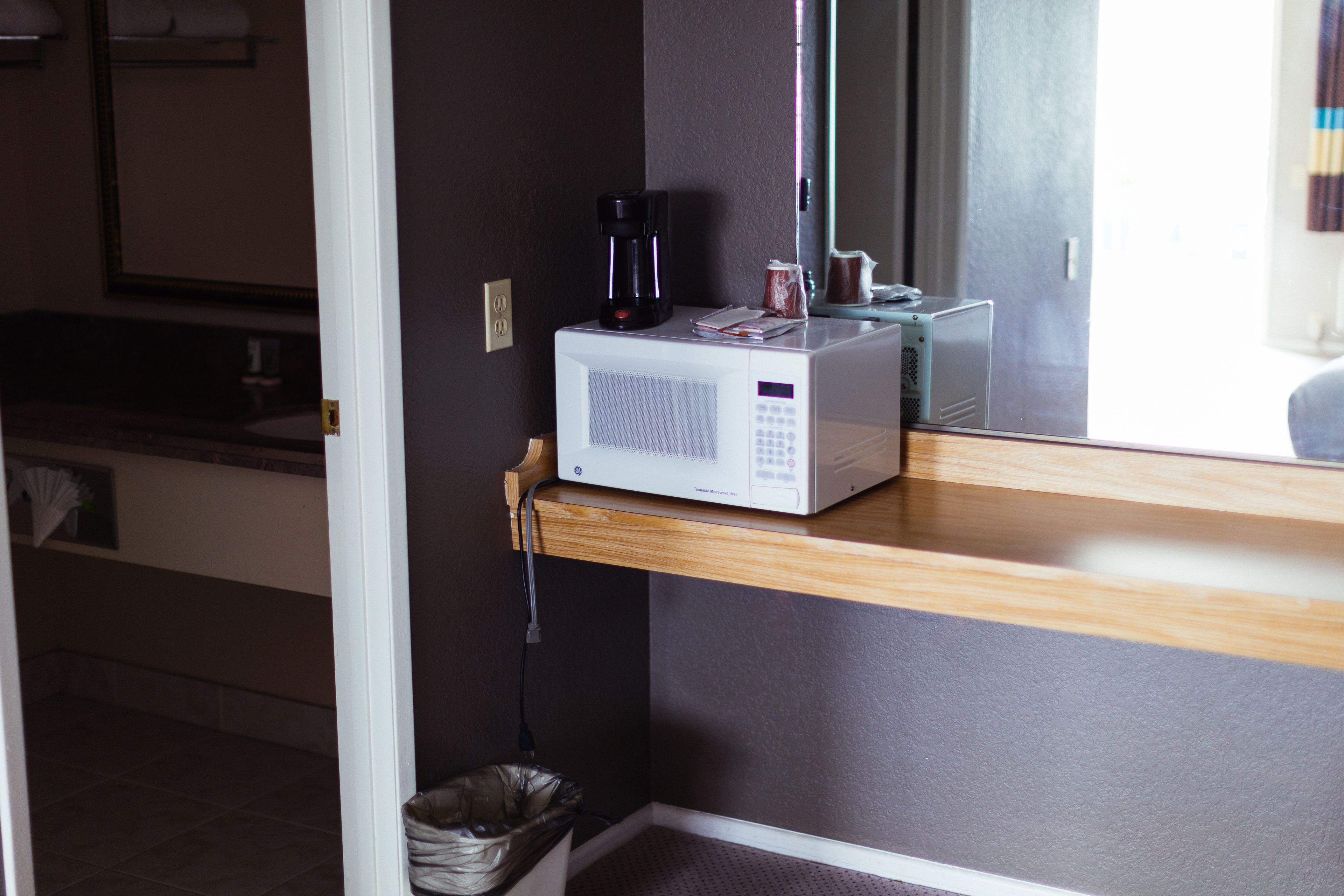 A microwave on a wooden shelf in a room with a dark wall and a window.