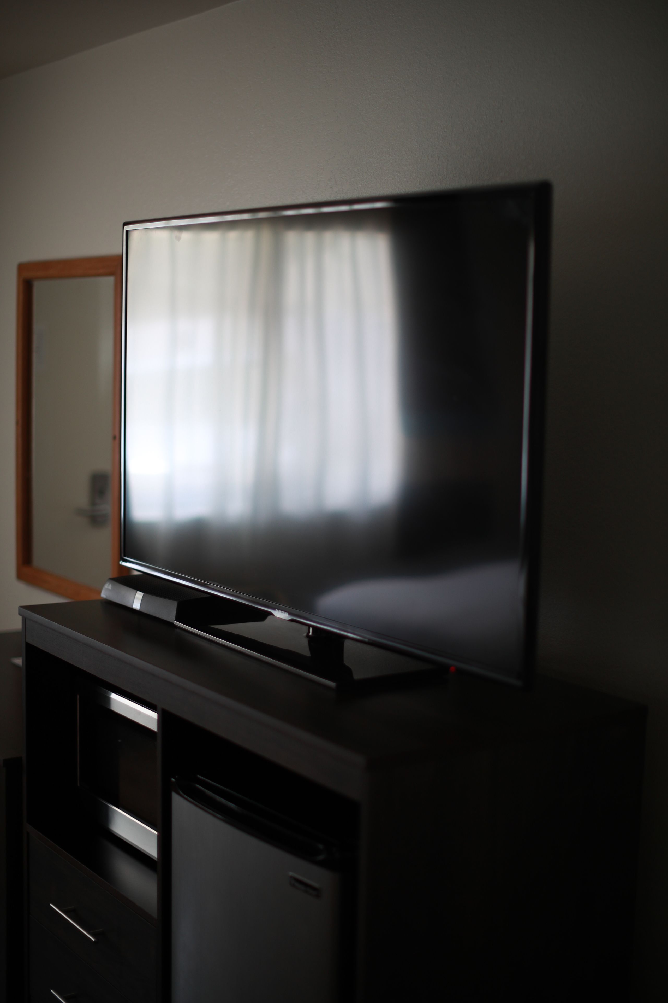 Un téléviseur à écran plat posé sur un meuble noir doté d’un cadre en bois, un petit réfrigérateur en dessous et un miroir accroché au mur.