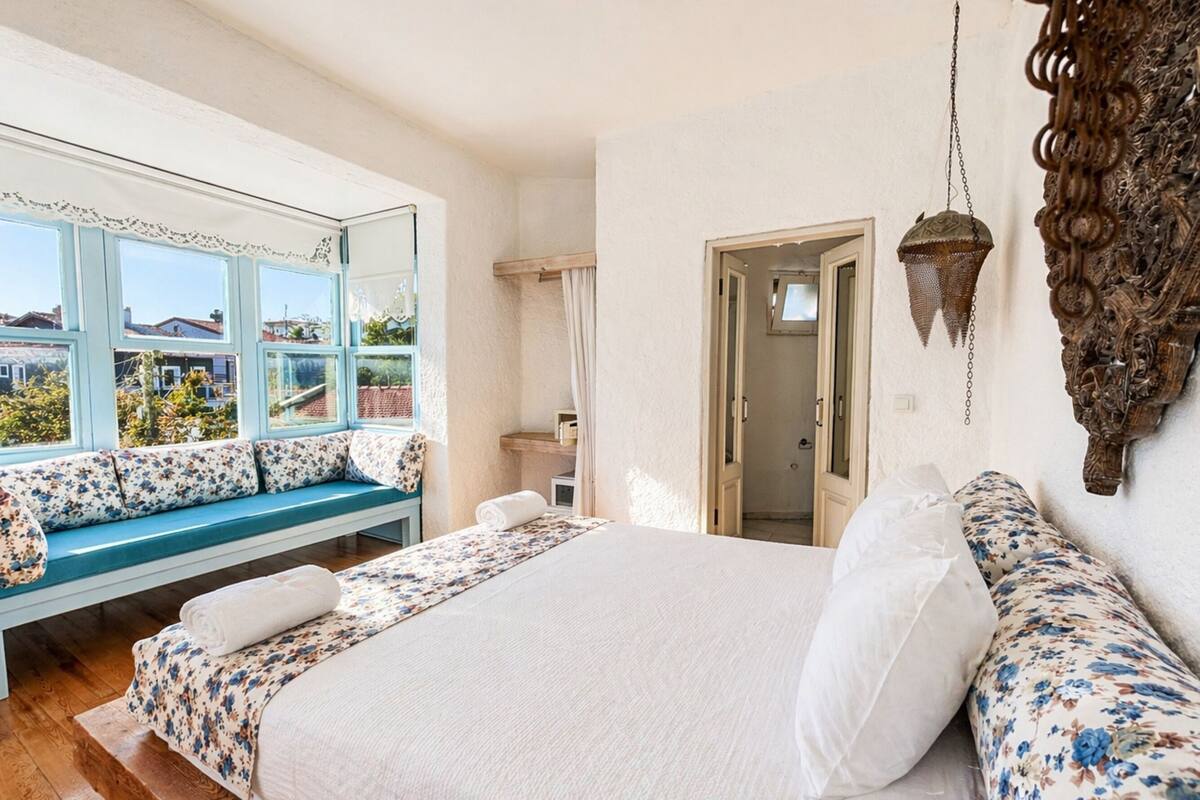 A bedroom with a large bed, a window seat, a mirror, and a door leading to a bathroom.