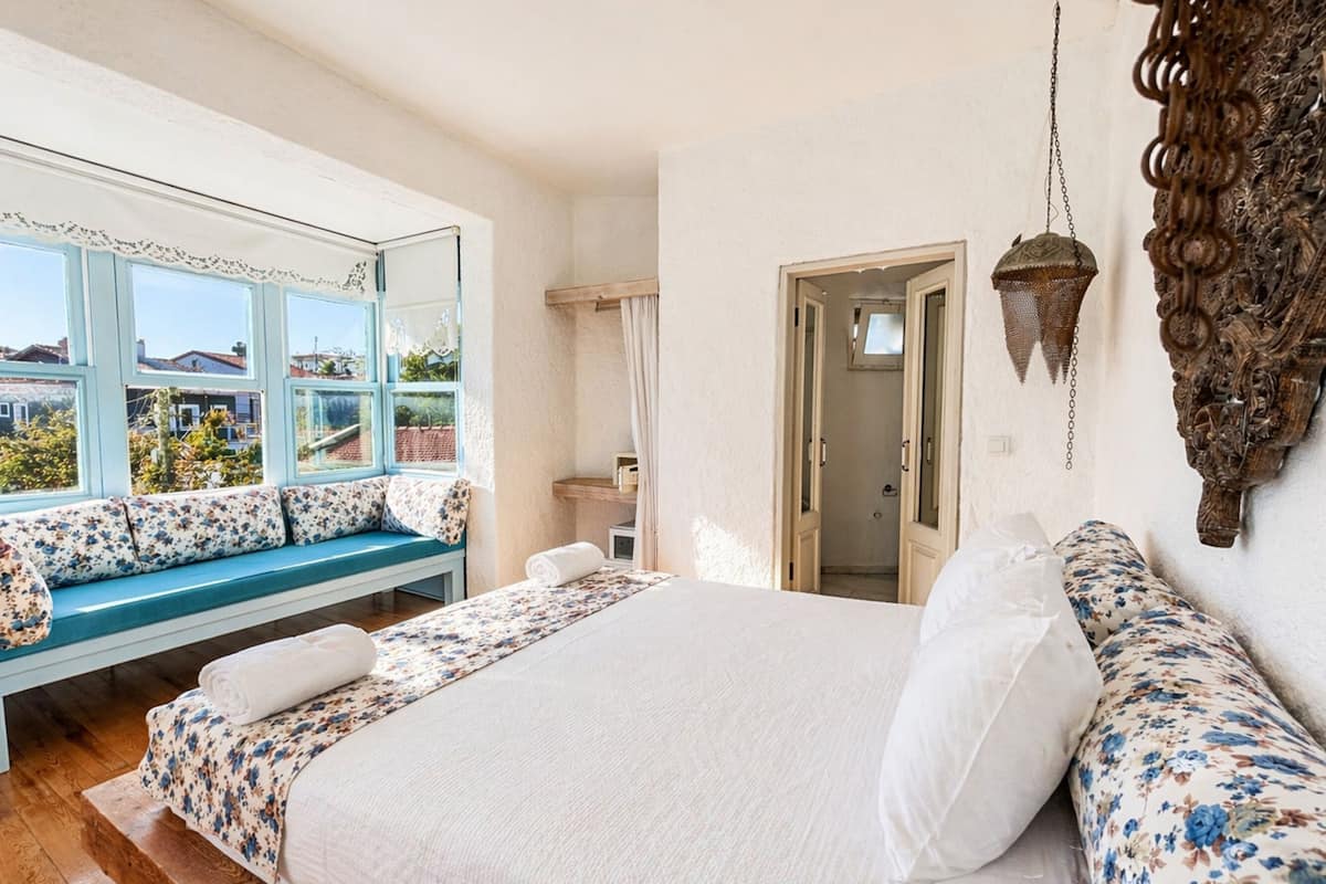 A bedroom with a large bed, a window seat, a mirror, and a door leading to a bathroom.