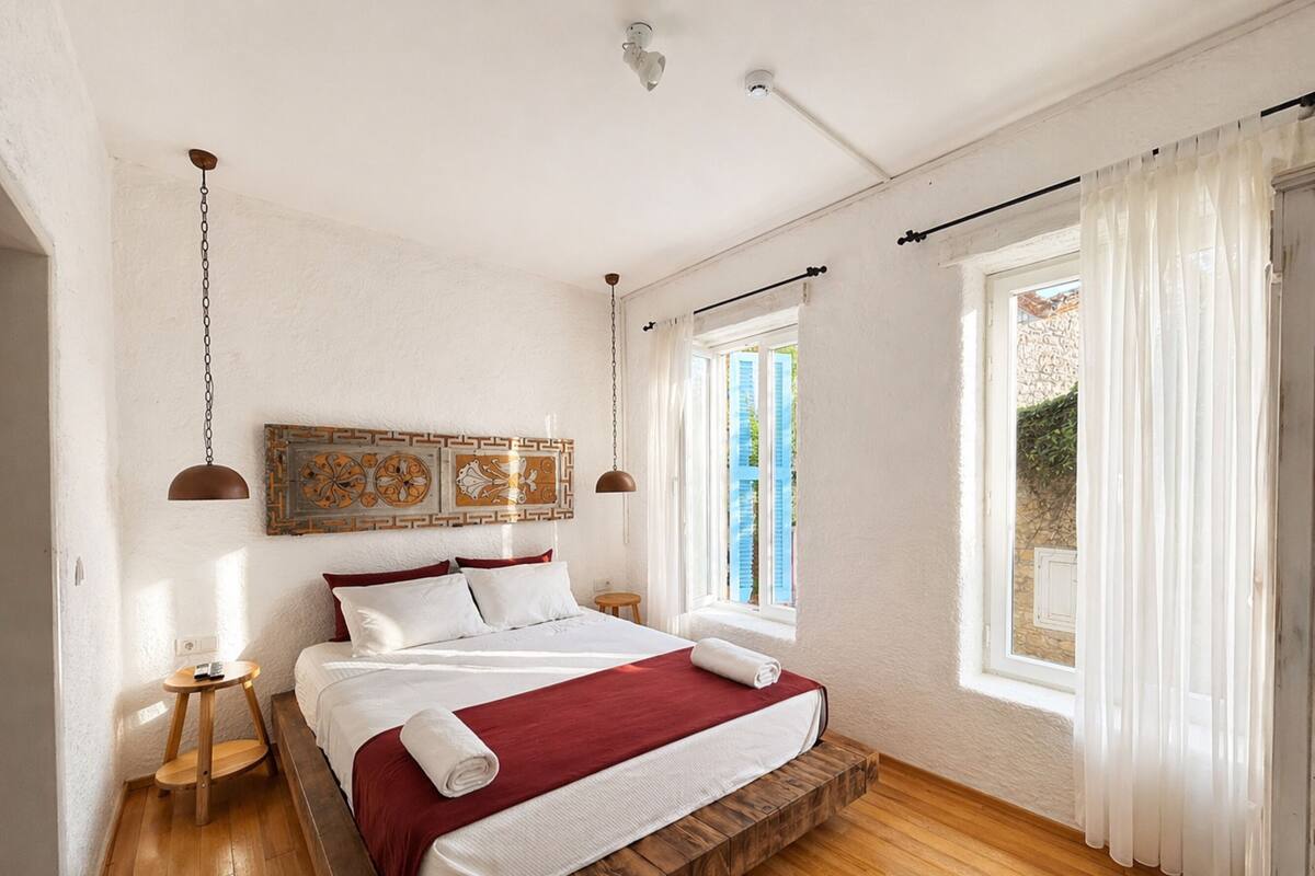 A bedroom with a wooden bed frame, white bedding, a red throw, a wooden bedside table, a wall-mounted light, and a window with sheer curtains.