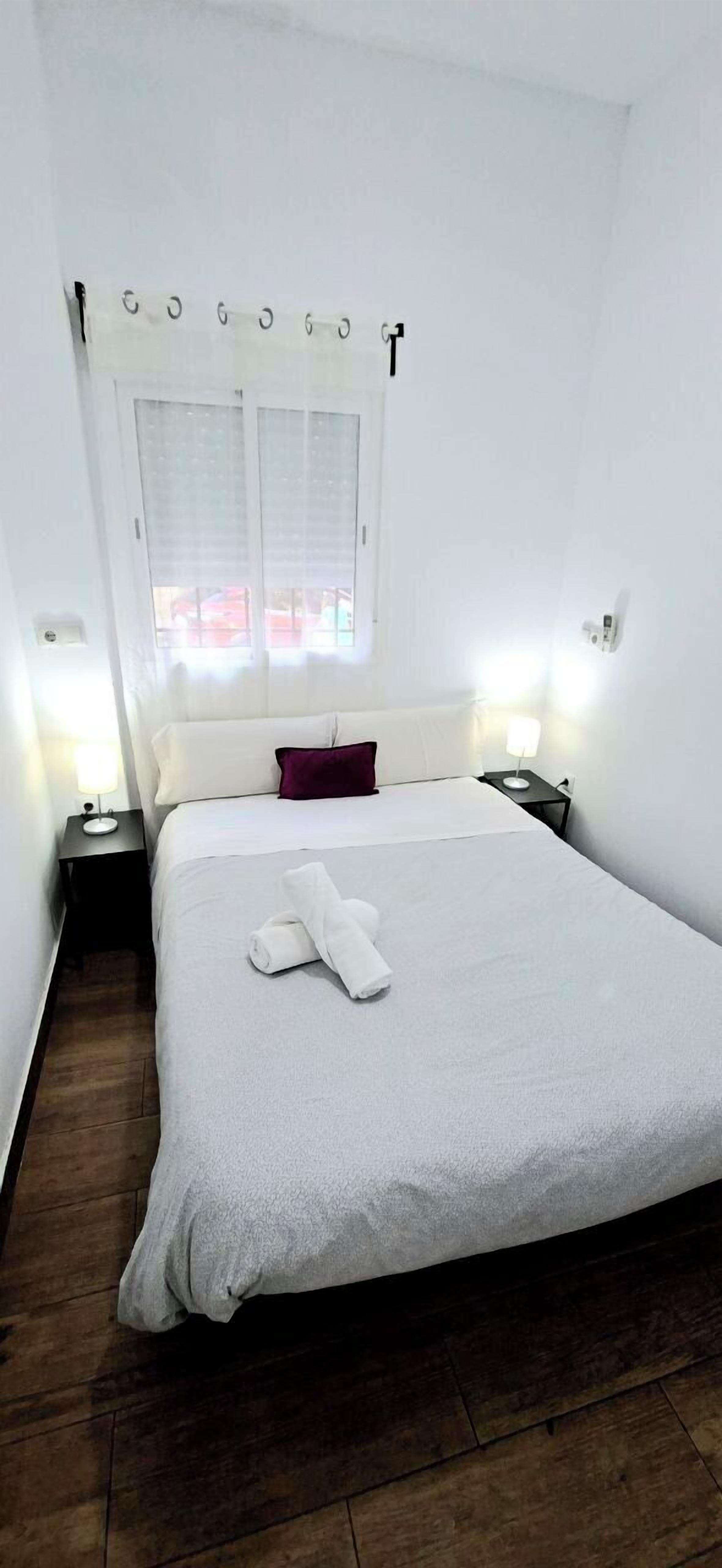 Una cama bien hecha con edredón blanco y una toalla enrollada, una ventana con persianas blancas, una mesita de noche con lámpara y una lámpara de pared.