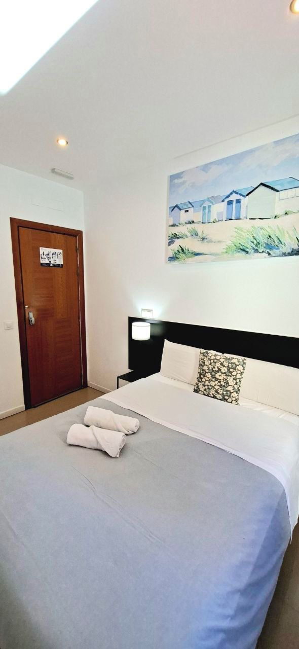 A neatly made bed with white linens and two rolled towels, a wooden door, and a painting of beach houses on the wall.