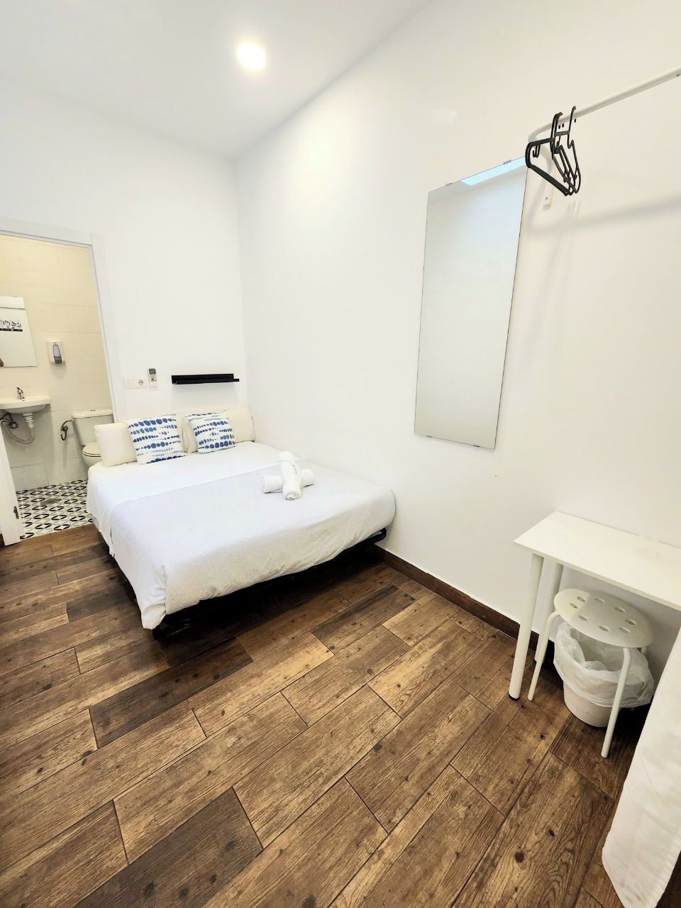 A modern hotel room with a wooden floor, a bed with white bedding, a small white desk, and a mirror on the wall.
