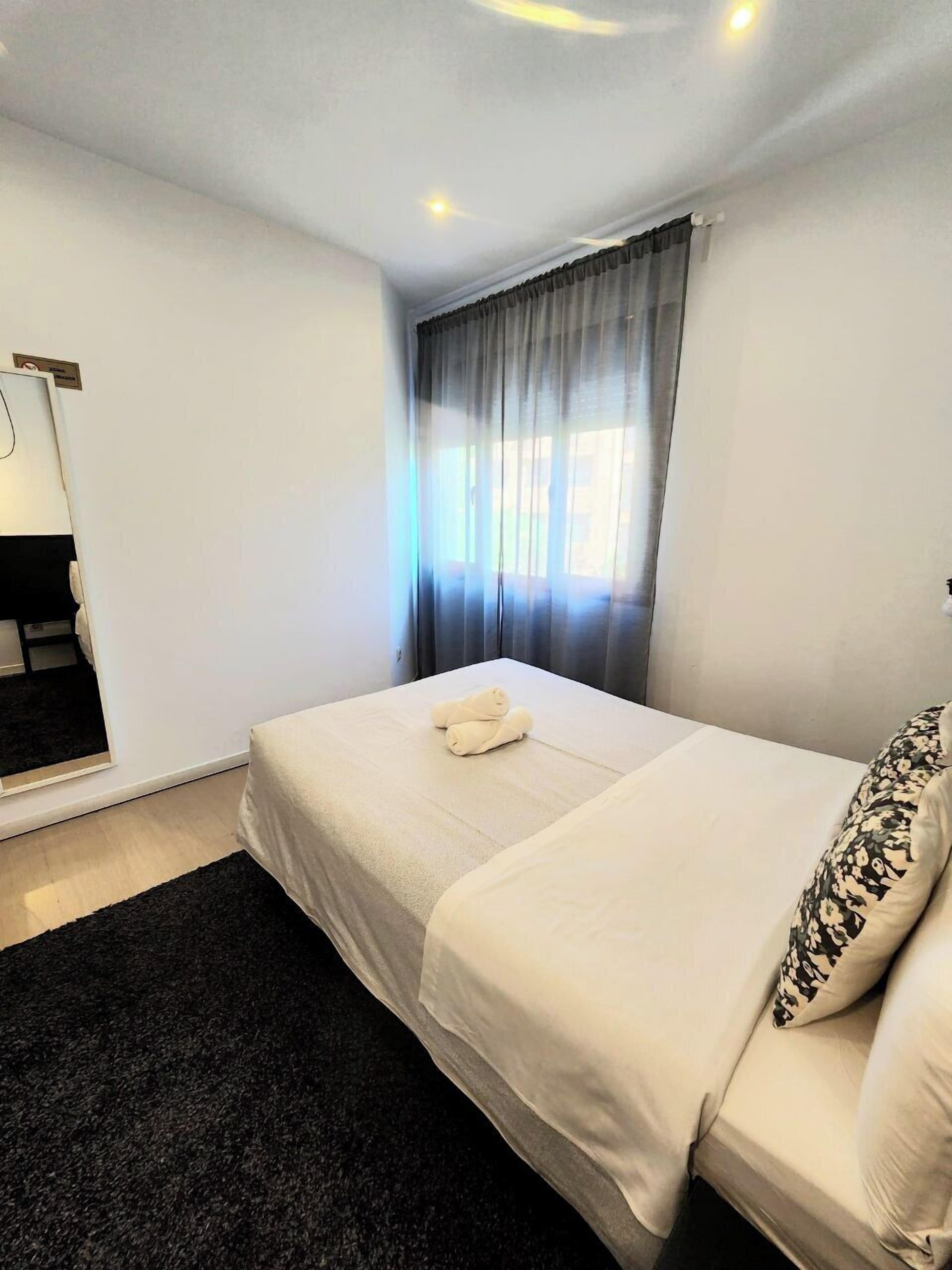 A hotel room with a bed, a black rug, a window with curtains, and a mirror on the wall.
