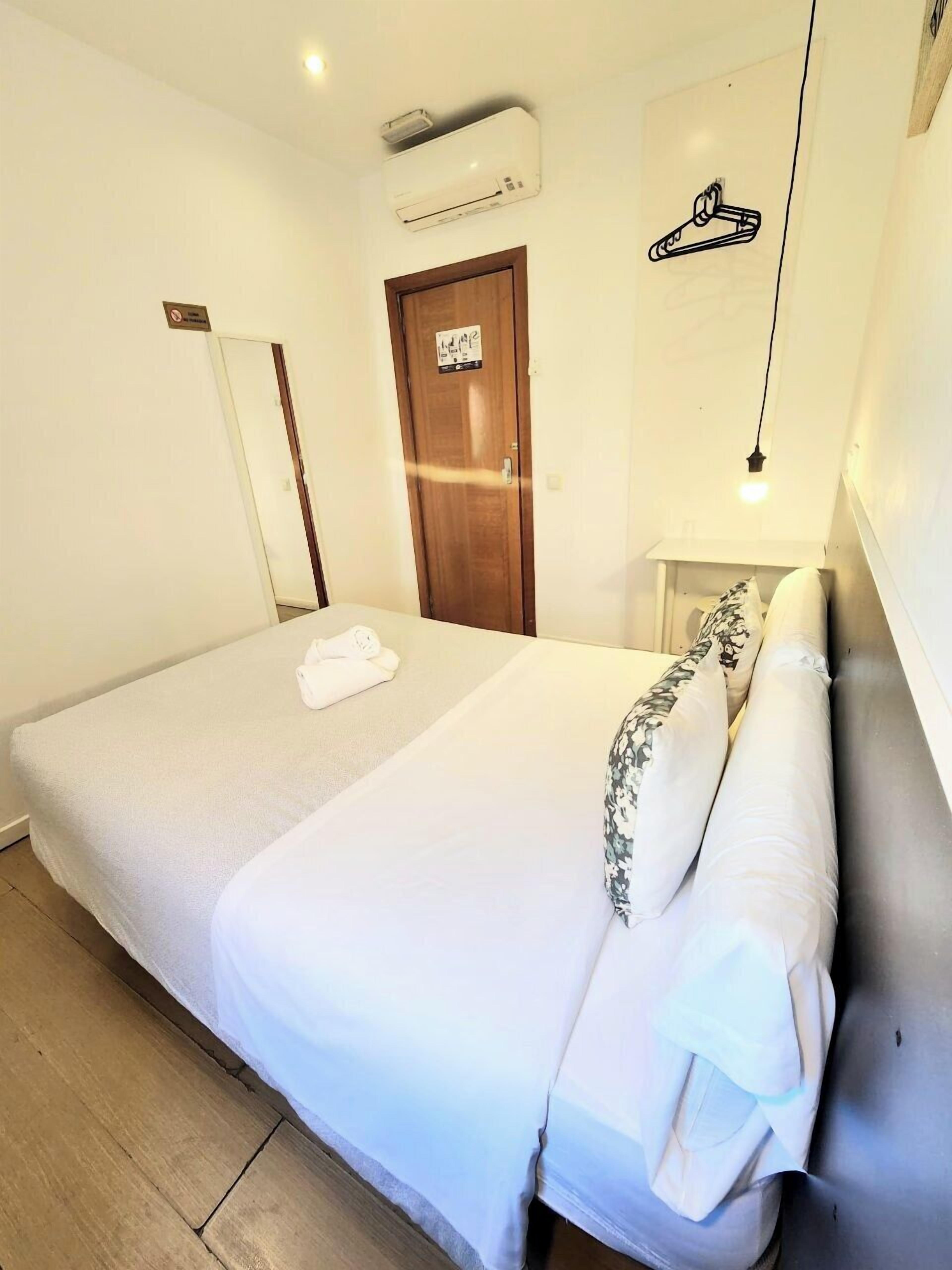 Una cama bien hecha con edredón blanco y almohadas florales, una puerta de madera con un cartel, una mesita de noche con una lámpara y un equipo de aire acondicionado en la pared.