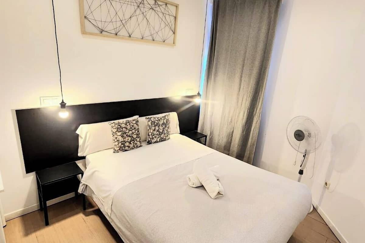 Una cama bien hecha con edredón blanco y almohadas, cabecera negra, mesita de noche pequeña, una obra de arte abstracto colgada en la pared, un ventilador de pie y una ventana con cortinas.