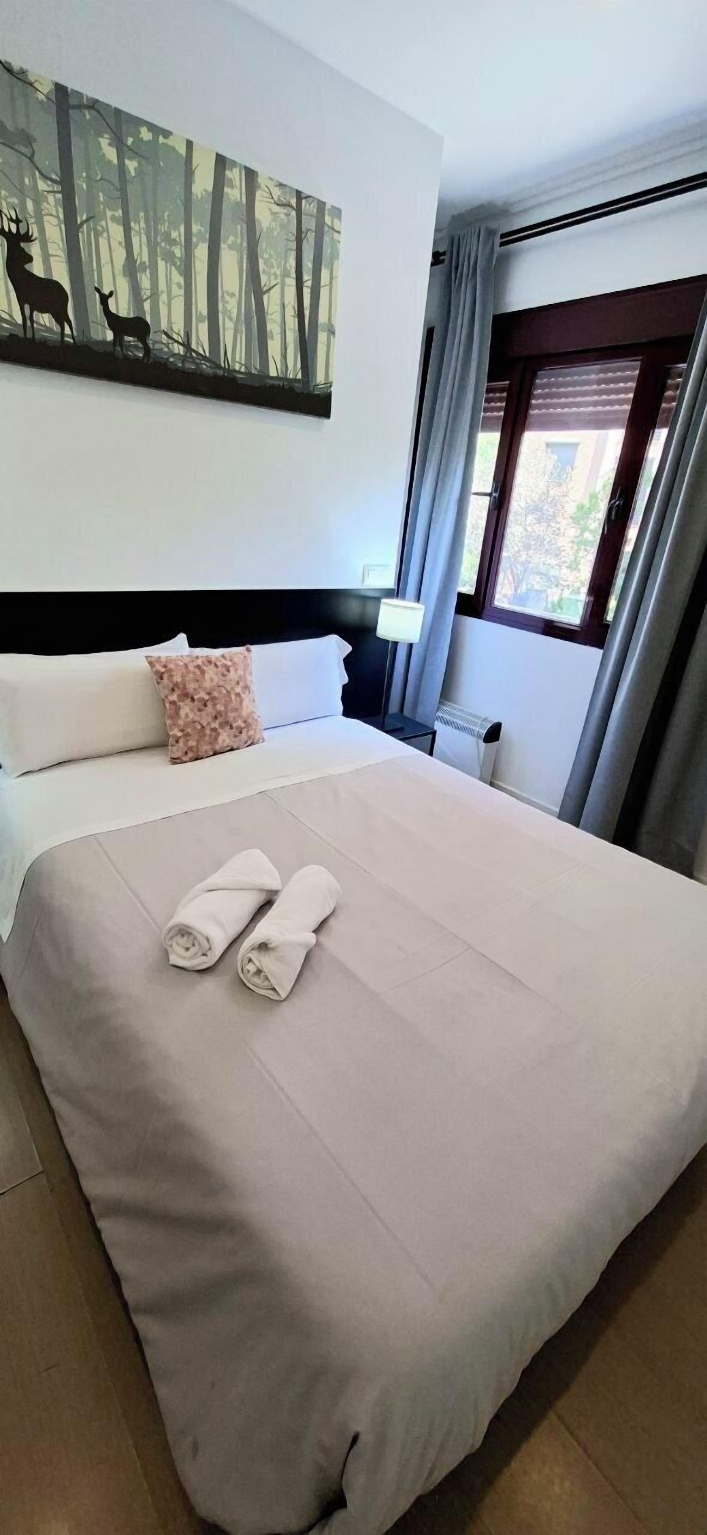 A neatly made bed with two rolled towels, a headboard, a nightstand with a lamp, a painting of deer on the wall, and a window with curtains.