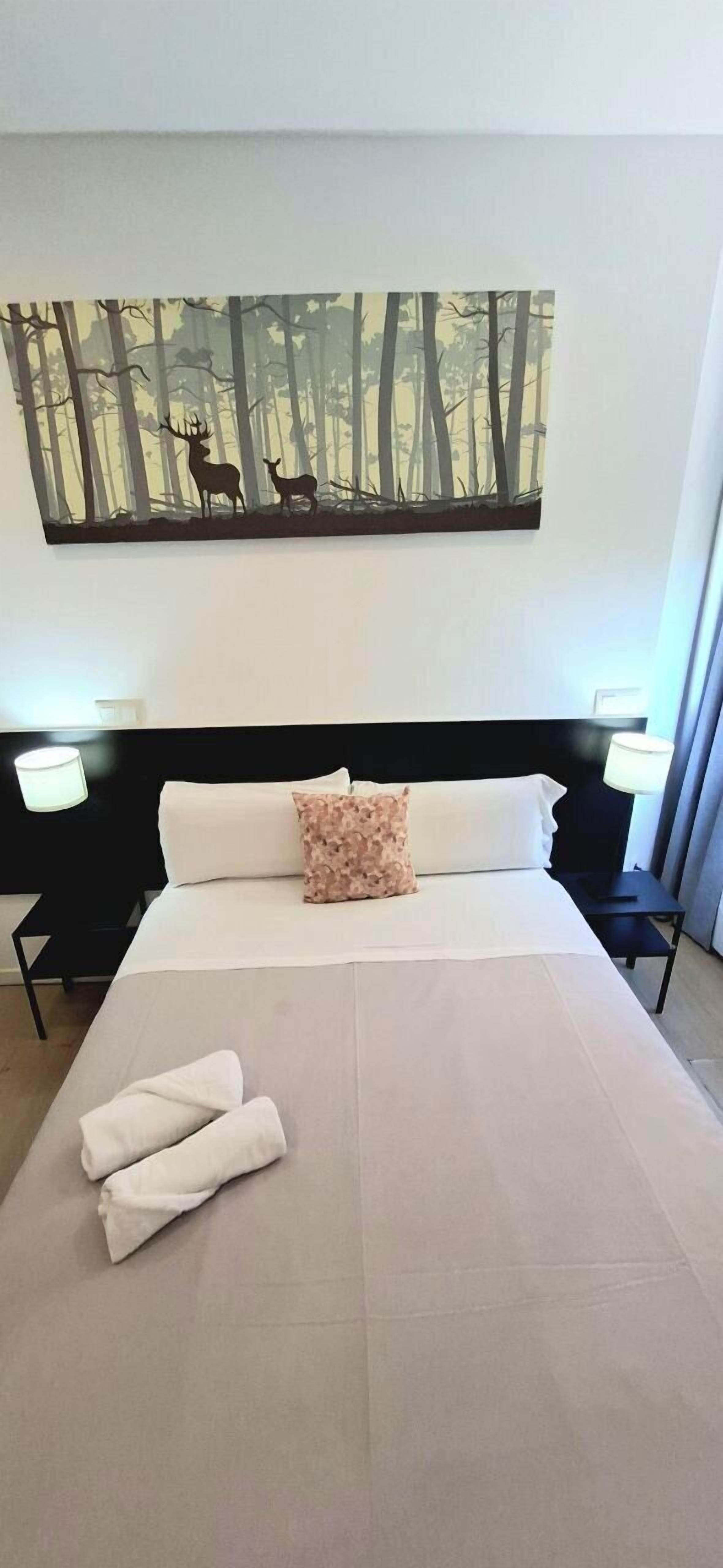 A neatly made bed with white linens and two rolled towels, a black headboard, and a forest-themed painting above it.
