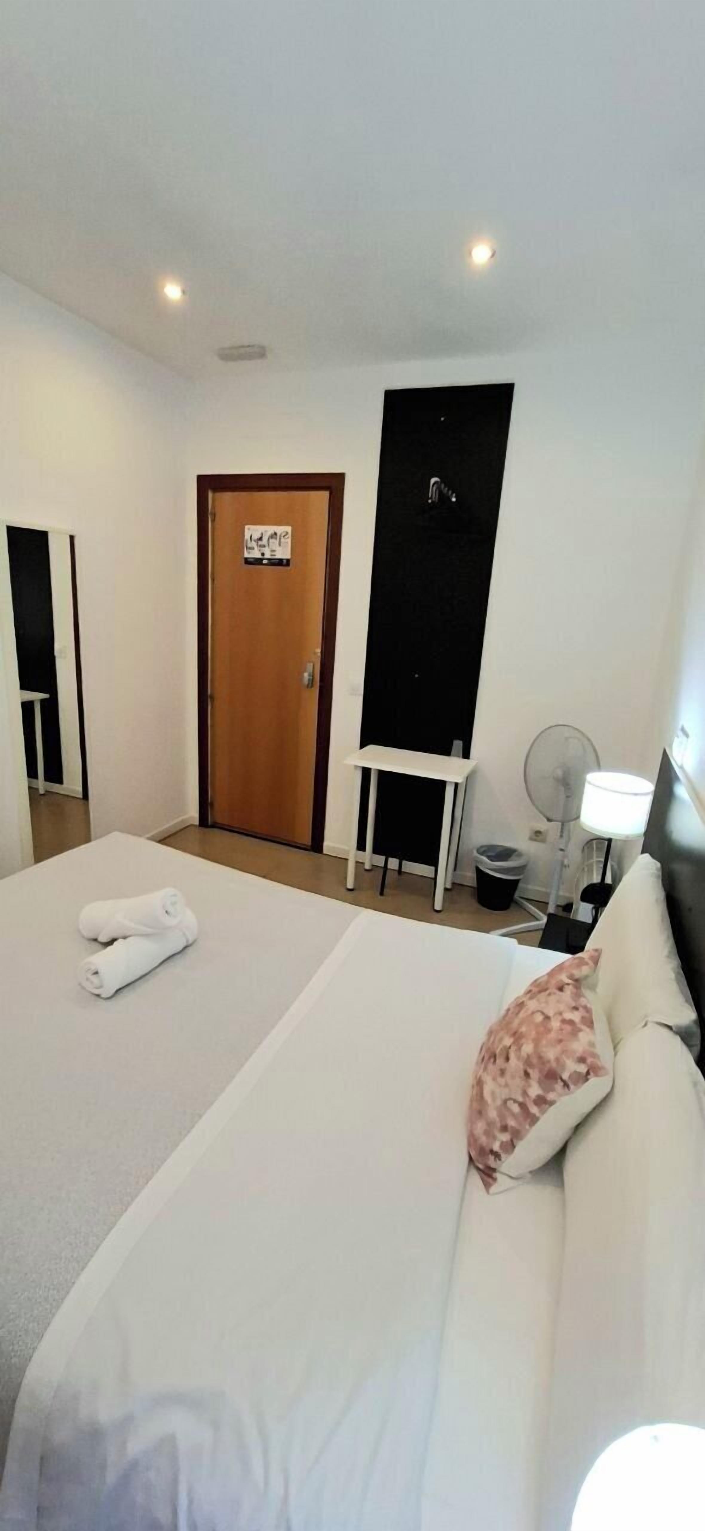 A hotel room with a bed, a desk, a chair, a lamp, and a door.