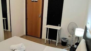 A hotel room with a bed, a desk, a chair, a lamp, and a door.