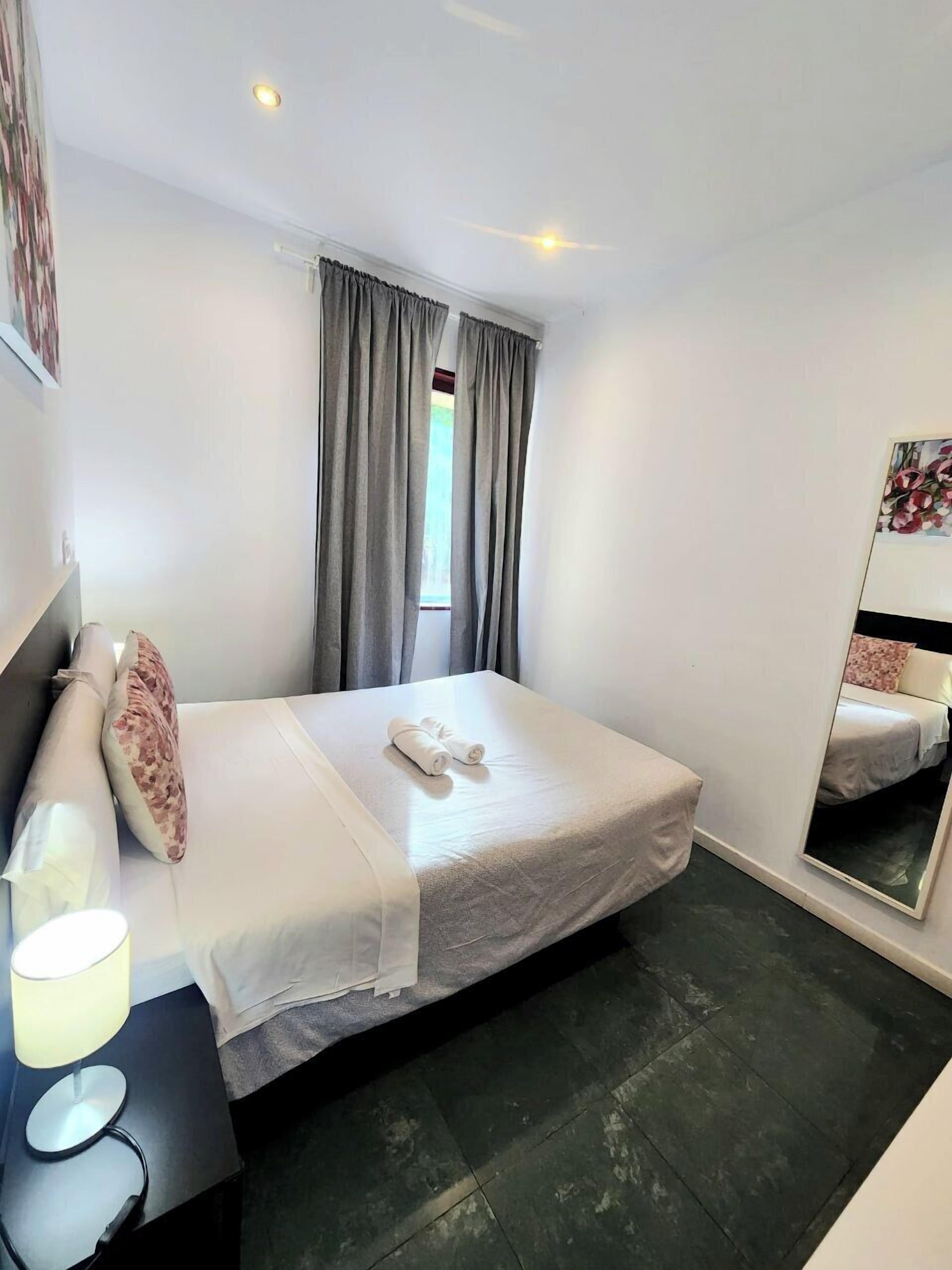 Et hotelværelse med en enkelt seng, et natbord med lampe, et vindue med gardiner og et spejl på væggen.