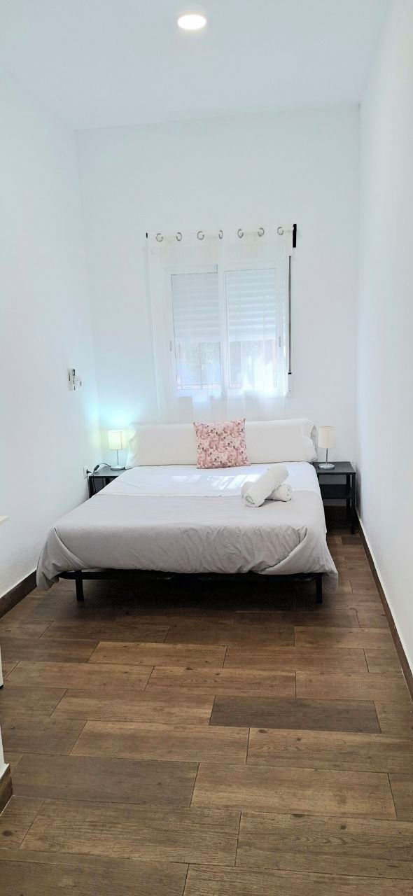 A bedroom with a bed, a nightstand, a window with blinds, and a wooden floor.