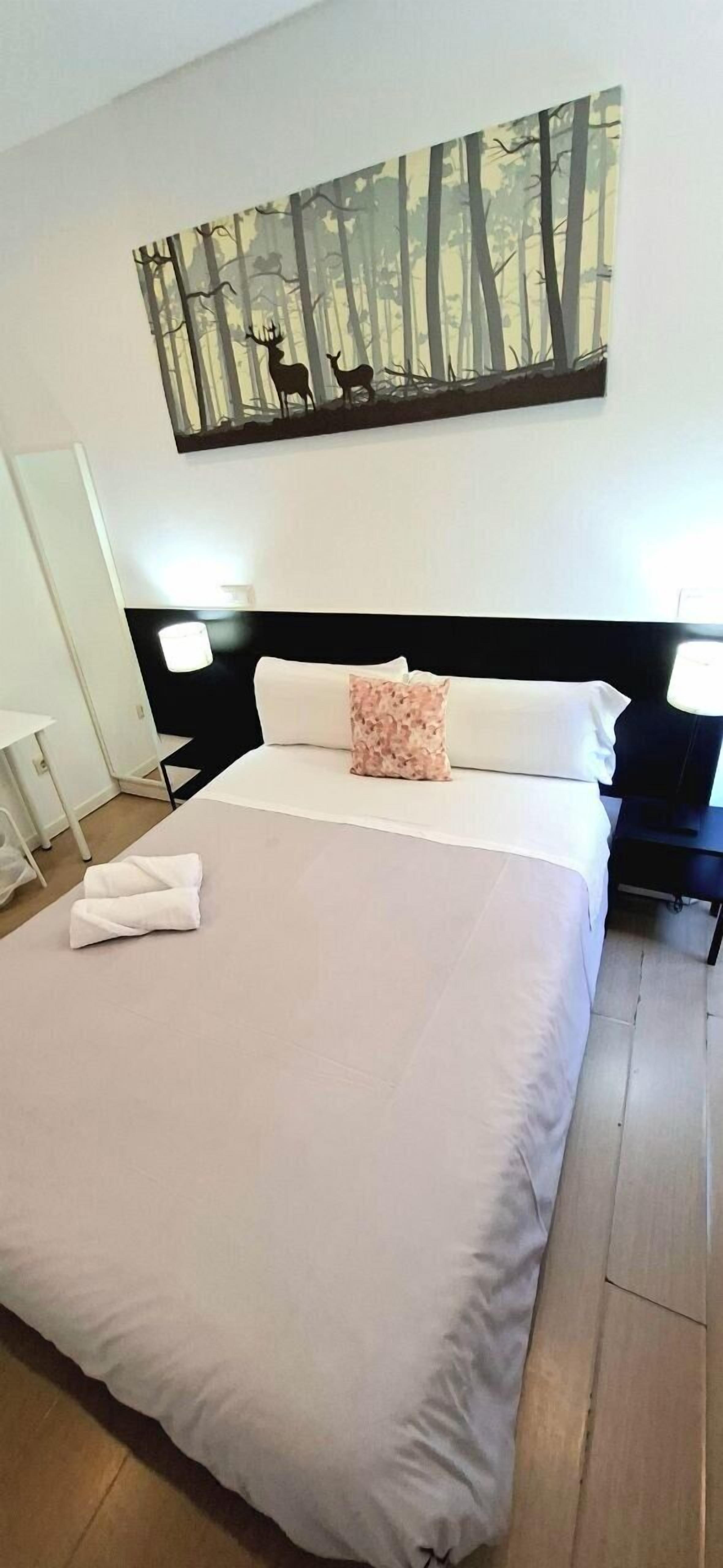 Una cama bien hecha con un cojín blanco y rosa, un cuadro de un bosque con ciervos en la pared, y una mesita de noche con una lámpara.